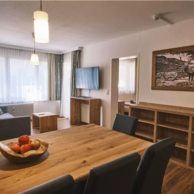 A modern living room with wood finishes and a large window front. On the dining table, there is a bowl of fresh apples.