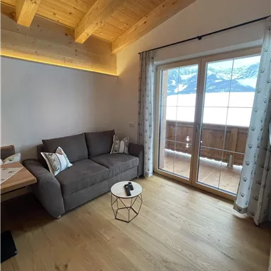 A cozy living room with a gray sofa and a small table. Through large windows, there is a view of the mountains and a balcony.