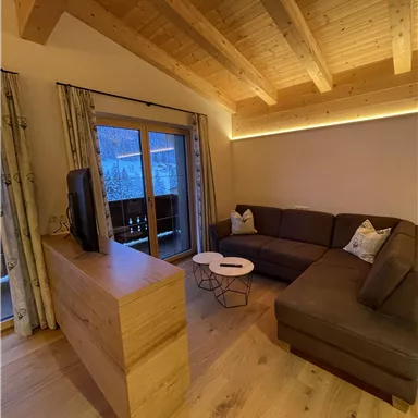 A modern living room with wooden ceilings and a cozy couch. Large windows offer a view of nature and provide plenty of daylight.
