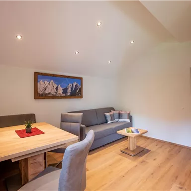 A cozy living space with wooden floors, a dining table, and a modern couch. A television hangs on the wall, along with a picture featuring mountain motifs.