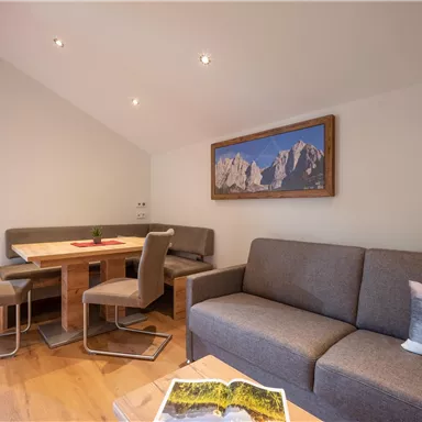 A modern living room with a gray sofa and a dining table. A landscape picture hangs on the wall, and there are windows with curtains.