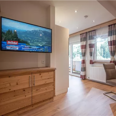 A modern living room with wooden surfaces and a large TV. Bright curtains and a dining table create an inviting atmosphere.