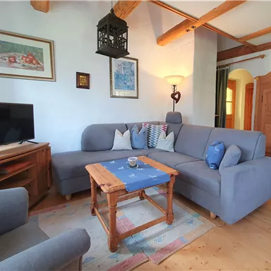 A cozy living room with a gray couch and a wooden table. Pictures are hanging on the wall, and a television can be seen in the background.