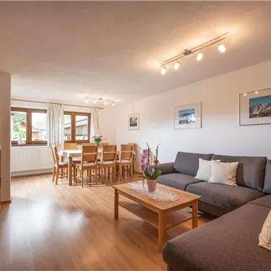 A cozy living room with a gray couch and a wooden table. In the background, there is a dining area with a dining table and chairs.