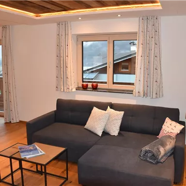 A modern living room with a comfortable sofa, a coffee table, and large windows. The wooden details and curtains give the room a cozy atmosphere.