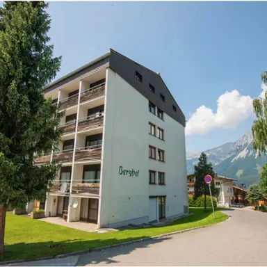 Ein modernes Gebäude mit mehreren Etagen und Balkonen, umgeben von grünen Bäumen. Im Hintergrund sind Berge und ein klarer blauer Himmel zu sehen.