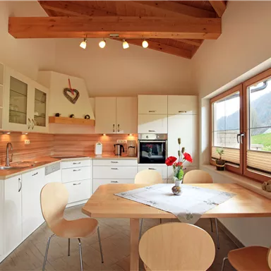 A modern kitchenette with white cabinets and wooden details. A round table with chairs sits in front of large windows that offer a view of the green landscape.