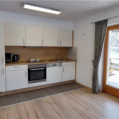 A modern kitchen with a stove, a refrigerator, and a seating area. Large windows provide plenty of daylight and a view of the snowy landscape outside.