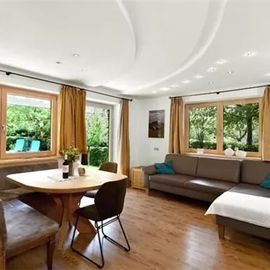 Ein helles Wohnzimmer mit einem gemütlichen Sofa und einem Essbereich. Große Fenster bieten Ausblick auf den Garten.