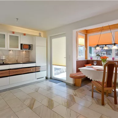 A modern kitchen with white cabinets and a dining area. Large windows let in plenty of light and there are orange curtains.