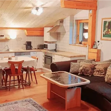 Eine gemütliche Wohnküche mit Holzdecke und moderner Ausstattung. Der Raum ist hell und einladend, mit einem Sofa und einem Esstisch für vier Personen.