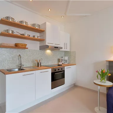 Eine moderne, helle Küche mit weißen Schränken und Holzregalen ist sichtbar. Es gibt einen kleinen Tisch mit einer Vase voller Blumen und eine lila Couch im Hintergrund.