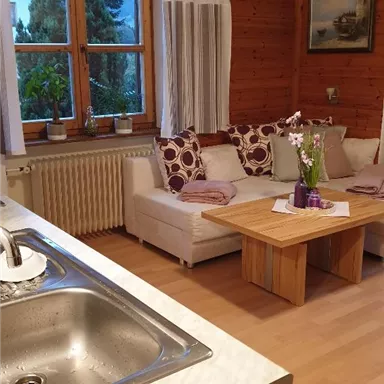 A cozy living room with a white couch and colorful cushions. The windows offer a view of greenery, and the wooden walls give a warm, inviting impression.
