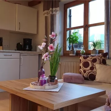 A cozy living room with a modern coffee table and beautiful cushions. Flowers and decorative elements are on the table, while large windows let in plenty of light.