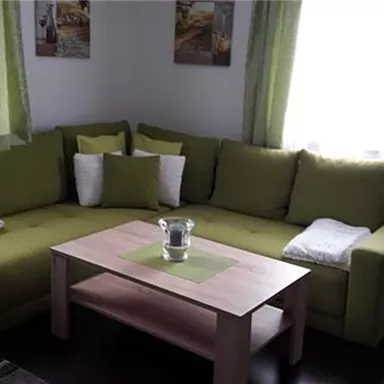 A cozy living room corner with a green sofa and cushions. A wooden table stands in the middle, surrounded by bright curtains and pictures on the wall.
