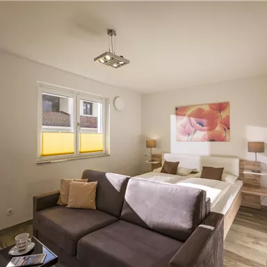 A modern bedroom with a large bed and a cozy sofa. Bright walls and natural light create an inviting atmosphere.
