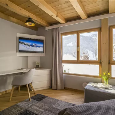 Ein modernes Schlafzimmer mit Holzbalkendecke und großen Fenstern. Der Raum ist hell und freundlich, mit einem Schreibtisch und einem Fernseher.