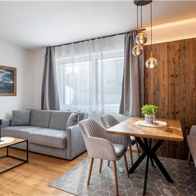 A modern living room with a gray sofa and a wooden table. Large windows provide plenty of daylight and a pleasant atmosphere.