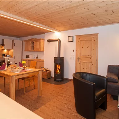 Ein gemütlicher Wohnraum mit Holzdecke und einem modernen Kamin. Eine Essecke und eine Küche sind ebenfalls sichtbar, die warme Atmosphäre lädt zum Entspannen ein.