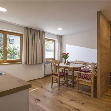 A cozy kitchen with a small dining area and large windows. The floor is made of wood and there are fresh flowers on the table.