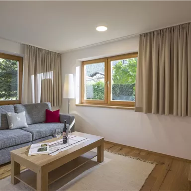 A cozy living room with a gray sofa and red pillows. Large windows let in a lot of light and offer a view of the greenery.