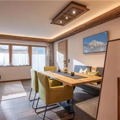 Ein modernes Wohnzimmer mit einem Esstisch und grünen Stühlen. Die Fenster bieten Tageslicht und einen Blick auf eine Berglandschaft.