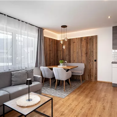 A modern living room with a gray sofa and a dining table for four people. Large windows let in plenty of light and create an inviting atmosphere.