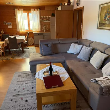 A cozy living room with a large gray couch and a wooden table. In the background, a dining table with chairs and a kitchen can be seen.