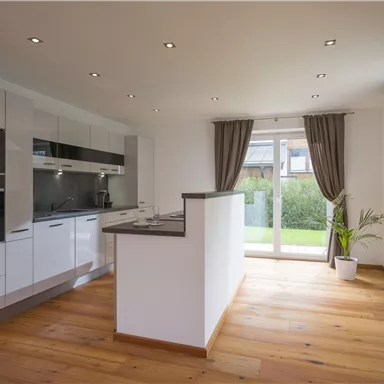 Eine moderne, helle Küche mit weißen Schränken und Holzfußboden. Große Fenster sorgen für viel Tageslicht und einen Blick in den Garten.