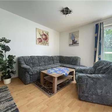 A cozy living room with a gray sofa set and a table. Large window with a view of greenery and decorative pictures on the wall.