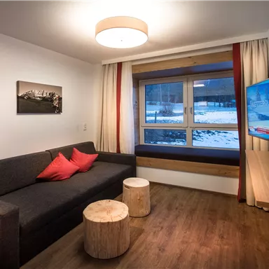 Ein modernes Wohnzimmer mit einem grauen Sofa und roten Kissen. Es gibt einen Fernseher und einen Blick auf eine schneebedeckte Landschaft durch das große Fenster.
