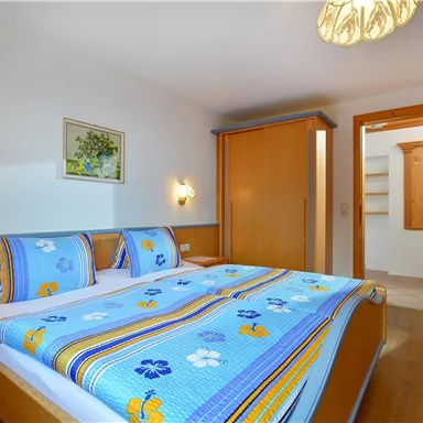 A cozy bedroom with a large double bed and colorful bedspreads. The room features warm wood surfaces and a bright, modern style.