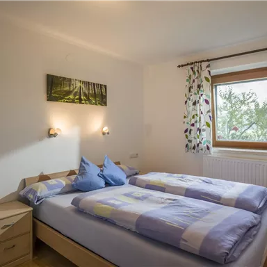 A cozy bedroom with two beds and colorful cushions. A window offers a view of the outdoors, and decorative curtains complement the space.