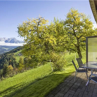 A beautiful view of the mountains with a green hill and colorful trees. A table with chairs is on a terrace.