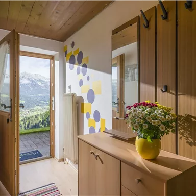 An inviting hallway with wood paneling and an open entrance. On a piece of furniture stands a vase with colorful flowers, and the mountains are visible in the background.