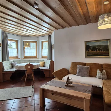 Ein gemütliches Wohnzimmer mit Holzbalkendecke und großen Fenstern. Der Raum ist mit einer bequemen Couch, einem Tisch und rustikalen Dekorationen eingerichtet.