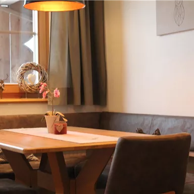 A cozy dining area with a wooden table and a bench. A decorative piece hangs on the wall and a flower serves as table decoration.
