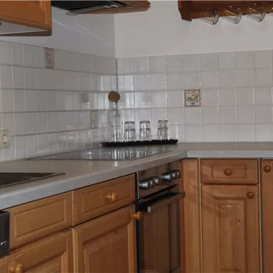 A bright kitchen with wooden cabinets and white tiles. It is equipped with a stove, a sink, and a microwave.