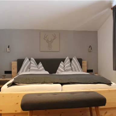 A cozy bedroom with a large double bed and modern wooden furniture. A picture of a stag’s head hangs on the wall, and the room has large windows with curtains.