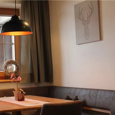 A cozy dining area with a wooden bench and a table. A modern lamp hangs over the table, and an antler-like artwork adorns the wall.
