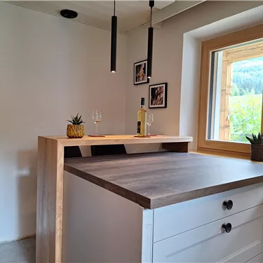 A modern kitchen corner with a wood bar. In the background, there are windows overlooking nature and some plants on the windowsill.