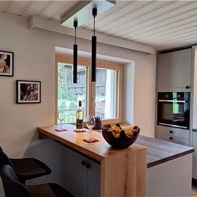 A modern kitchen with wooden surfaces and elegant bar stools. On the table, there is a fruit bowl and in the background, there are windows letting in natural light.