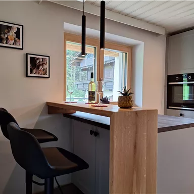 A modern kitchen with a wooden bar and two black chairs. Windows with a view and decorations on the walls.