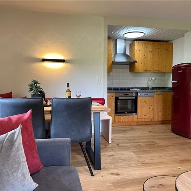 A modern living room with an open kitchen. The room is decorated in warm colors and features wooden furniture and comfortable seating.