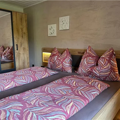 A cozy bedroom with two beds and colorful cushions. In the background, there is a wardrobe with a mirror and there are light-colored walls.