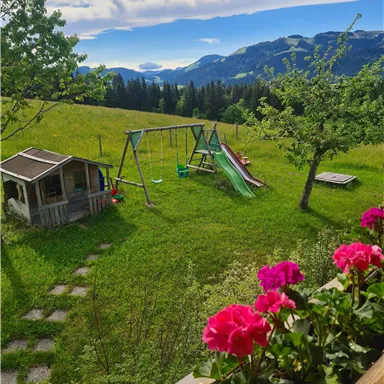 Eine grüne Wiese mit Spielgeräten und einem kleinen Spielhaus. Im Vordergrund blühen pinke Blumen und im Hintergrund sind Berge sichtbar.