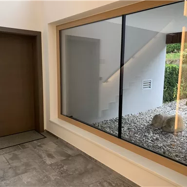 A modern entrance area with a large glass panel and a view of a small garden. The walls are light and the door is made of wood.