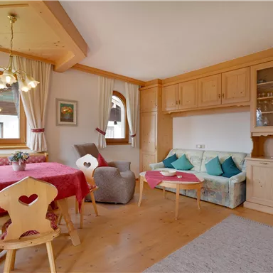 A cozy living room with a large table, chairs, and a light wooden decor. There is a sofa area with cushions and large windows that let in plenty of light.