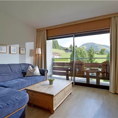 Ein helles Wohnzimmer mit einem blauen Sofa und einem Holztisch. Große Fenster bieten einen Blick auf die Berge und die grüne Landschaft draußen.