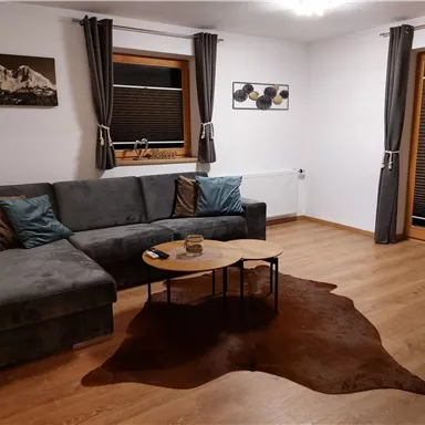 Ein gemütliches Wohnzimmer mit einem grauen Sofa und einem runden Tisch. Große Fenster sorgen für einen hellen Raum und die Holzfußböden verleihen eine warme Atmosphäre.